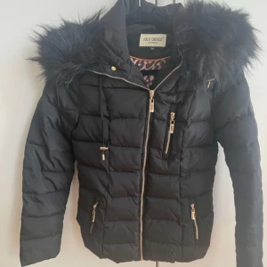 Svart pufferjacka Roco Baroco med fuskpäls - Svart pufferjacka från Roco Baroco med snygg fuskpäls på huvan och guldfärgade dragkedjor. Jackan har flera fickor med dragkedja och quiltad design. Perfekt för kalla dagar och ger en cool vibe till din outfit. I bra skick!