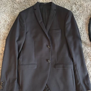 Svart kavaj tiger of sweden - Stilren svart kavaj med klassisk passform och två knappar framtill. Kavajen har smala slag, tre knappar vid ärmslut och två fickor med lock samt en bröstficka. Perfekt för en dressad look.