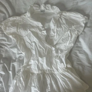 Vit omlottblus från Hunkydory - Supersöt vit blus från Hunkydory Essentials i 100% bomull. Blusen har v-ringning, volangdetaljer och korta ärmar. Perfekt för varma dagar och ger en fräsch och luftig känsla.