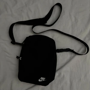 Svart axelväska från Nike - Säljer en stilren svart axelväska från Nike med justerbar rem och klassisk vit logga framtill. Väskan har dragkedja och är perfekt för att bära med sig det viktigaste. Enkel och clean design som funkar till det mesta.