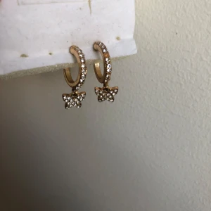 Guldiga örhängen med glittriga fjärilar - Supersöta guldiga örhängen med små gnistrande stenar och hängande fjärilar. Perfekta för att ge din look lite extra bling och söt vibe. Små hoops med glittriga detaljer som verkligen fångar ljuset.