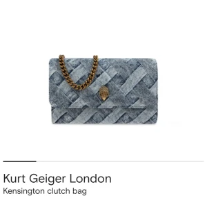 Kurt Geiger väska - Snygg clutch från Kurt Geiger i blått jeanstyg med flätat mönster. Väskan har en grov guldfärgad kedja och ikonisk örndetalj framtill. Perfekt storlek för det viktigaste och ger en cool touch till din outfit. Går ner i pris vid snabb affär