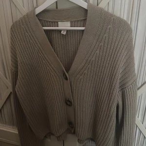 Beige ribbstickad kofta med knappar - Superfin beige ribbstickad kofta med stora knappar framtill och v-ringad hals. Modellen är croppad och har långa ärmar. Perfekt att slänga över ett linne eller t-shirt.