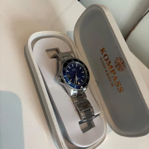 Blå-silver klocka Kompass Stockholm - Stilren klocka från Kompass Stockholm med blå urtavla och datumvisning. Klockan har en robust länk i silverfärgat metall och tydliga indexmarkeringar. Perfekt accessoar för dig som gillar klassisk design med modern touch.