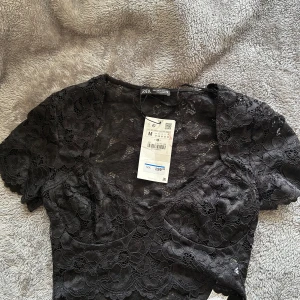 Svart spetsblus från Zara - Supersnygg svart blus från Zara i genombruten spets med korta ärmar och v-ringning. Croppad modell med fina vågiga kanter och blommigt spetsmönster. Perfekt att styla med höga jeans eller kjol för en trendig look.