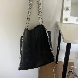 Svart axelväska med kedjerem - Snygg svart axelväska i skinnimitation med grova silverfärgade kedjor som axelrem. Väskan har en stilren och modern design med mjuk struktur och kedjedetaljer längs kanterna. Perfekt för dig som gillar en edgy och trendig look.