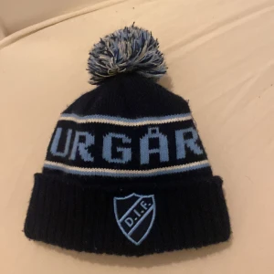 Djurgården mössa  - Mörkblå och ljusblå stickad mössa från Djurgårdens IF med stor tofs på toppen. Mössan har broderad DIF-logga framtill och texten 'DJURGÅRDEN' runt om. Perfekt för att visa ditt lagstöd under kalla dagar.