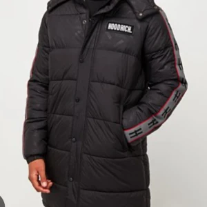 Svart lång dunjacka från Hoodrich - Lång svart dunjacka från Hoodrich med huva och logga på bröstet. Jackan har quiltad design, knappar och dragkedja framtill samt gråa detaljer med svarta symboler och röd kant längs ärmarna. Perfekt för kalla dagar och riktigt snygg streetstil.