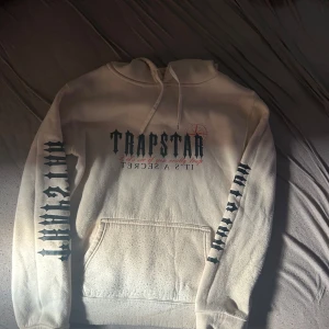  Beige trapstar hoodie - Säljer en vit hoodie från Trapstar med svart tryck på bröstet och båda ärmarna. Hoodien har huva med snörning och en stor magficka. Materialet är mjukt och skönt, perfekt för chill dagar. Snygg streetwear-vibe och coola detaljer. 