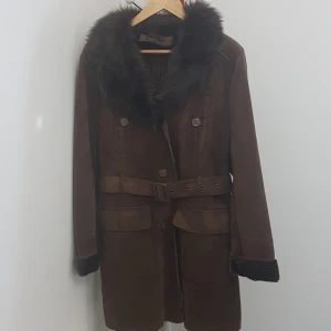 Brun mocka kappa med fuskpäls - Snygg brun kappa i mocka-look med bred krage i fuskpäls och bälte i midjan. Knappar framtill, stora fickor och pälsdetaljer vid ärmslut. Perfekt för kalla dagar när du vill ha en retro och cool vibe.