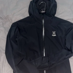 Svart Haglöfs jacka - Storlek ”M” . Skriv dm för prisförslag samt video! Säljer en svart vindjacka från Haglöfs med huva och hel dragkedja framtill. Jackan har diskret logga på bröstet, två sidofickor och meshfoder på insidan. Perfekt för blåsiga dagar och outdoor-aktiviteter. Enkel, stilren och funktionell design.