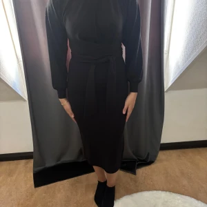 Svart långklänning med knytband - Stilren svart långklänning med långa ärmar och markerad midja tack vare ett brett knytband. Klänningen har en figurnära passform och är tillverkad i ett mjukt, stretchigt material. Perfekt för dig som gillar en klassisk och elegant look.