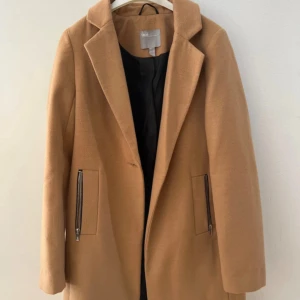 Beige kappa från ASOS, stl 36 - Snygg beige kappa från ASOS i klassisk rak modell med krage och långa ärmar. Jackan har två fickor med dragkedja och är fodrad på insidan. Perfekt för dig som vill ha en stilren och tidlös look.