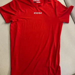 Röd CCM t-shirt i polyester - Röd t-shirt från CCM med vit logga på bröstet. Klassisk rund halsringning och korta ärmar. Tillverkad i mjuk och lätt polyester som andas, perfekt för sport eller chill. Enkel och clean design utan mönster.