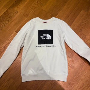 Vit sweatshirt från The North Face - Säljer en stilren vit sweatshirt från The North Face med svart logga och texten 'Never Stop Exploring' på bröstet. Tröjan har rund hals, långa ärmar och ribbade muddar. Perfekt för dig som gillar enkel och clean streetstyle.