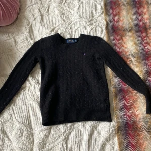 Svart kabelstickad tröja Polo Ralph Lauren - Svart kabelstickad tröja från Polo Ralph Lauren i storlek XS. Klassisk modell med rund halsringning och lila broderad logga på bröstet. Tröjan har långa ärmar och ribbade muddar. Perfekt för dig som gillar tidlös och stilren design.❤️