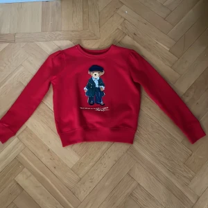 Röd Polo Bear tröja från Ralph Lauren - Superfin röd tröja från Ralph Lauren med Polo Bear-tryck på bröstet. Klassisk rund halsringning och långa ärmar med lätt puff vid axlarna. Mjuk insida och ribbade muddar. Perfekt för dig som gillar statement-plagg med ikonisk design.❤️ ALDRIG använd❤️