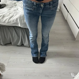 Blå bootcut jeans med låg midja - Snygga blå jeans med bootcut passform och låg midja. Jeansen har klassisk femficksdesign, ljus tvätt och subtila slitningar för en avslappnad vibe. Tillverkade i mjukt denimtyg som sitter skönt hela dagen.