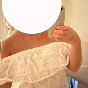 Vit offshoulder blus med volang - Superfin vit offshoulder blus med bred volang och broderade hålmönster. Toppen är ärmlös och har resår upptill för att sitta snyggt på plats. Perfekt för varma dagar och ger en romantisk vibe. Tillverkad i luftig bomull.