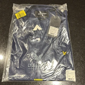 Marinblå skjorta från Lyle & Scott XL - Snygg marinblå skjorta från Lyle & Scott i slim fit-modell. Skjortan är tillverkad i bomullspoplin och har klassisk krage, långa ärmar och knäppning framtill. Ikonisk gul logga broderad på bröstet ger en stilren touch.