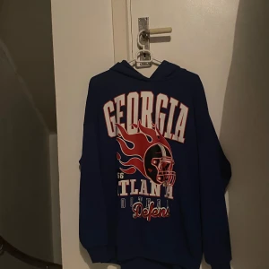 Blå hoodie GEORGIA från New yorker  - Blå hoodie från New yorker  med stor GEORGIA-tryck och coolt flames-motiv på framsidan. Tröjan har huva, långa ärmar och en avslappnad passform. 