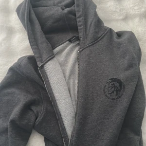 Vintage diesiel zip up - Säljer en mörkgrå hoodie från Diesel med dragkedja och huva. Tröjan har ett svart tryck med Diesel-logga på bröstet, två fickor framtill och är tillverkad i mjuk bomullsmix. Perfekt för chill dagar och streetwear-vibe.