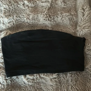 Svart bandeau tubtopp från 157 - Enkel och stilren svart bandeau tubtopp från 157. Toppen är axelbandslös och har en tight passform som sitter snyggt på kroppen. Perfekt att matcha med höga jeans eller kjol för en clean look.