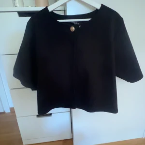 Svart kavajjacka med kort ärm - Stilren svart kavajjacka från Missäy med korta ärmar och rund halsringning. Jackan har en snygg guldig knapp framtill som stängning och är i en rak, boxig modell. Perfekt för dig som gillar minimalistisk och clean stil.