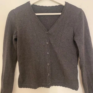 Mörkgrå kofta med v-ringning - Mysig mörkgrå kofta med v-ringning och knappar hela vägen framtill. Långärmad modell med ribbade kanter och diskret vågig kant längst ner. Lappen är tyvärr avklippt men passar en som är xs 