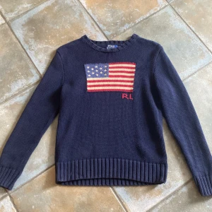 Mörkblå stickad tröja Polo Ralph Lauren - Mörkblå stickad tröja från Polo Ralph Lauren med amerikansk flagga och RL-broderi på bröstet. Tröjan har rund hals, ribbade muddar och lång ärm. Perfekt för dig som gillar klassisk och stilren design.
