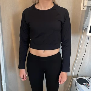 Svart croppad tröja från New Apparel - Säljer en svart, croppad långärmad tröja från New Apparel. Tröjan är ribbad och har en tight passform med rund halsringning. Tillverkad i stretchigt material av polyamid och spandex, perfekt för träning eller chill. Diskret logga på bröstet.