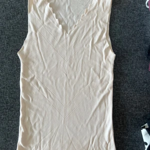 Beige ribbad ärmlös linnetopp - Snygg beige linnetopp med ribbad struktur och v-ringad hals med vågig kant. 