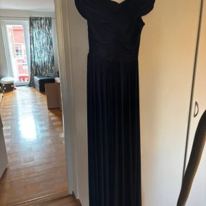 Marinblå långklänning offshoulder - Säljer en elegant marinblå långklänning med offshoulder-modell och v-ringning. Klänningen har rynkade detaljer över bysten och ett mjukt, fallande tyg som ger en snygg siluett. Perfekt för bal eller festliga tillfällen.