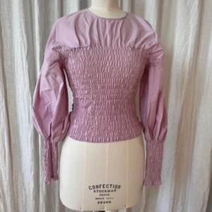 Ljuslila blus med puffärmar - Supersöt ljuslila blus med smockad midja och långa puffärmar. Blusen har rund halsringning och smockdetaljer vid ärmslut och överkropp för en riktigt trendig look. Perfekt för dig som gillar romantiska och feminina plagg.