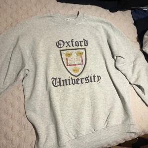 Grå Oxford University VINTAGE sweatshirt - Snygg grå sweatshirt med Oxford University-tryck och klassiskt vapensköldmotiv framtill. Tröjan har rund halsringning och långa ärmar, perfekt för en avslappnad och stilren look. Passar dig som gillar college-vibe och enkel stil.