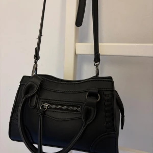 Svart axelväska med dragkedja - Snygg svart axelväska i skinnimitation med både handtag och justerbar axelrem. Väskan har en dragkedja framtill och coola detaljer längs sidorna. Perfekt storlek för det viktigaste och passar till många olika outfits.💓