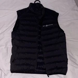 Svart dunväst från Sail Racing - Svart dunväst från Sail Racing med quiltad design och hög krage. Västen har dragkedja framtill och två sidofickor med dragkedja. Logotyp på bröstet och i nacken. Perfekt för lager-på-lager och stilren look.