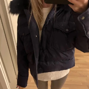 Mörkblå dunjacka från Meotine S/M - Säljer en mörkblå dunjacka från Meotine i storlek S/M. Jackan har fluffig fuskpäls på huvan, stora fickor med knappar och en riktigt snygg, boxig passform. Perfekt för kalla dagar när du vill vara både varm och stilren.
