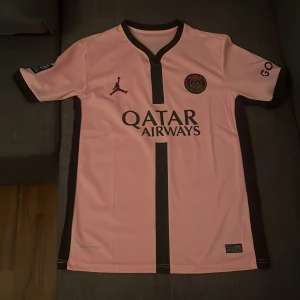 PSG rosa fotbollströja Jordan x Nike - Snygg rosa PSG fotbollströja med svarta detaljer och logga från Jordan och Paris Saint-Germain. Tröjan har korta ärmar, svart V-ringning och Qatar Airways-tryck på bröstet. Tillverkad i lätt och ventilerande material, perfekt för match eller träning.