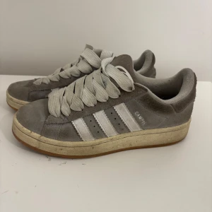 Grå Adidas Campus sneakers i mocka - Snygga Adidas Campus sneakers i grå mocka med klassiska vita ränder på sidorna och breda vita skosnören. Låg modell med gummisula och retrovibbar. Perfekta för dig som gillar streetstyle och tidlös design. Skorna är endast smutsiga och går att tvätta. 