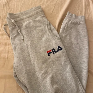 Grå mjukisbyxor från FILA - Snygga ljusgrå mjukisbyxor från FILA med logga på benet. Byxorna har ribbade muddar, dragsko i midjan.