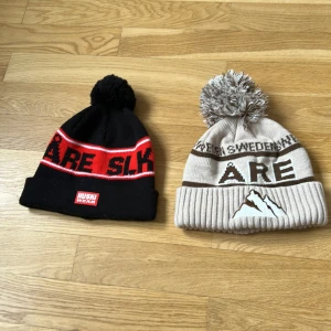 Två Åre-mössor med tofs - Två stickade Åre-mössor med tofs. Ena är svart och röd med texten 'ÅRE SLK' och Huski Wear-märke, den andra är beige med bruna detaljer, texten 'ÅRE' och bergmotiv. Båda har uppvikt kant och är perfekta för kalla dagar. Skickas på post, du står för frakt. Annars finns det tillgängligt i Östersund/åre. 1 för 60kr 2 för 100kr