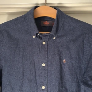 Blå skjorta Morris Slim Fit S - Snygg mörkblå skjorta från Morris i slim fit-modell. Skjortan har klassisk button down-krage, vita knappar och broderad logga på bröstet. Tillverkad i mjuk bomull, perfekt för en stilren look.