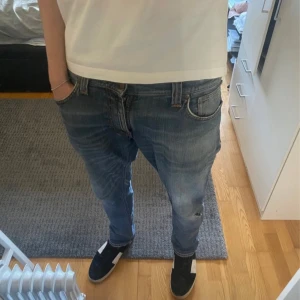 Blå Nudie Jeans, W30 L32 - Säljer ett par klassiska blå jeans från Nudie Jeans med snygga slitningar och orangea sömmar. Modellen har normal passform och raka ben, med ikoniska broderade bakfickor. Perfekta för dig som gillar en avslappnad men stilren look.