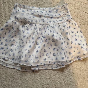 Vit blommig kortkjol med blått mönster - Supersöt vit kortkjol med blått blommönster och lätt volang. Kjolen har en bred midja och är gjord i ett tunt, luftigt material som passar perfekt till varma dagar. Den har en flowy känsla och ger en romantisk vibe.