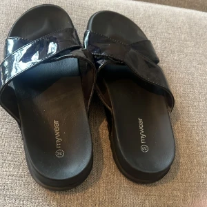 Svarta sandaler från mywear, strl 39 - Snygga svarta sandaler från mywear med breda, glansiga remmar i kors över foten. Platt sula och öppen tå ger en chill vibe. Perfekta för varma dagar när du vill ha något enkelt men ändå stilrent. Materialet är syntet med lackad finish.
