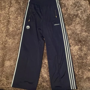 Adidas blå träningsbyxor wide fit - Mörkblå träningsbyxor från Adidas med klassiska vita ränder längs sidorna, broderad logga och Est. 1972-märke. Byxorna har bred passform, elastisk midja och dragkedjor på fickorna. Perfekta för en sportig och avslappnad stil.