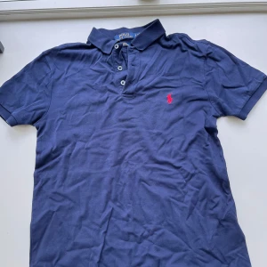 Marinblå pikétröja från Ralph Lauren - Snygg marinblå pikétröja från Ralph Lauren med klassisk krage, tre knappar och röd broderad logga på bröstet. Slim fit-modell i stretchig bomullsmix som sitter tajt och bekvämt. Perfekt för en clean och sportig look.