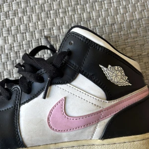 Nike Air Jordan 1 Mid svart/vit/rosa - Snygga Nike Air Jordan 1 Mid sneakers i svart och vitt med rosa swoosh. Klassisk siluett med svart snörning, rund tå och vit sula. 