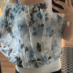 Blommig vit och blå blus - Luftig blus från Culture med stora blå och svarta blommor på vit botten. Modellen är lös och har bred resår i nederkant och halsringning. Tunt, transparent tyg som ger en fräsch och somrig vibe. Perfekt till lågmidjade jeans eller en söt kjol.